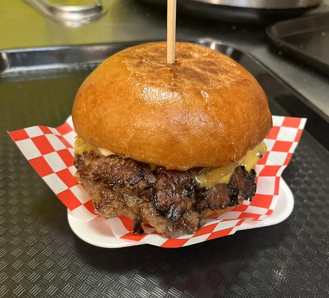 Smash burger with a crispy-edged patty and melted cheese on a brioche bun with a wooden skewer, on red and white checkered paper