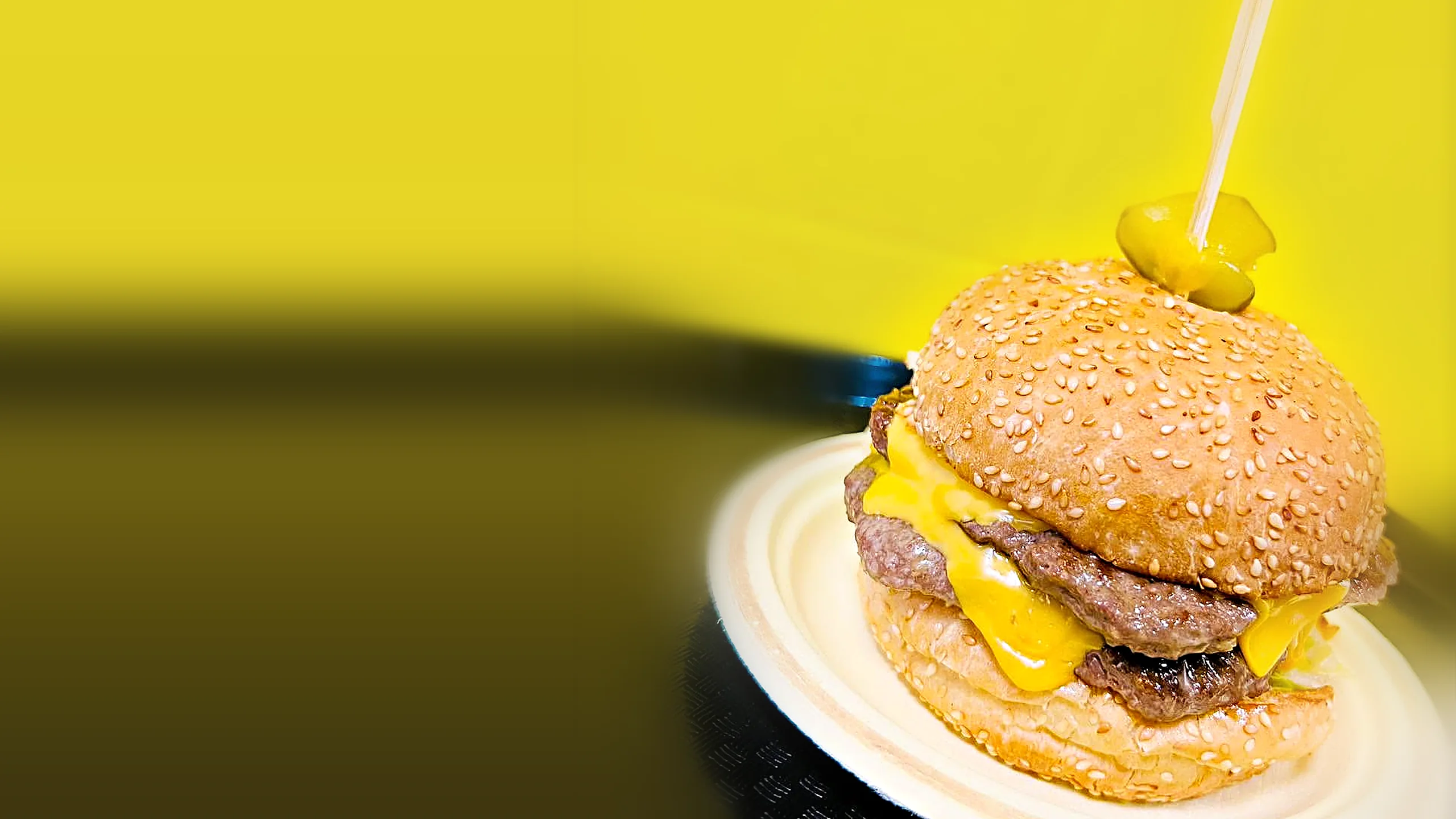 Double Oklahoma smash burger with two beef patties, melted cheddar and a pickle on a skewer, on a paper plate against a yellow wall